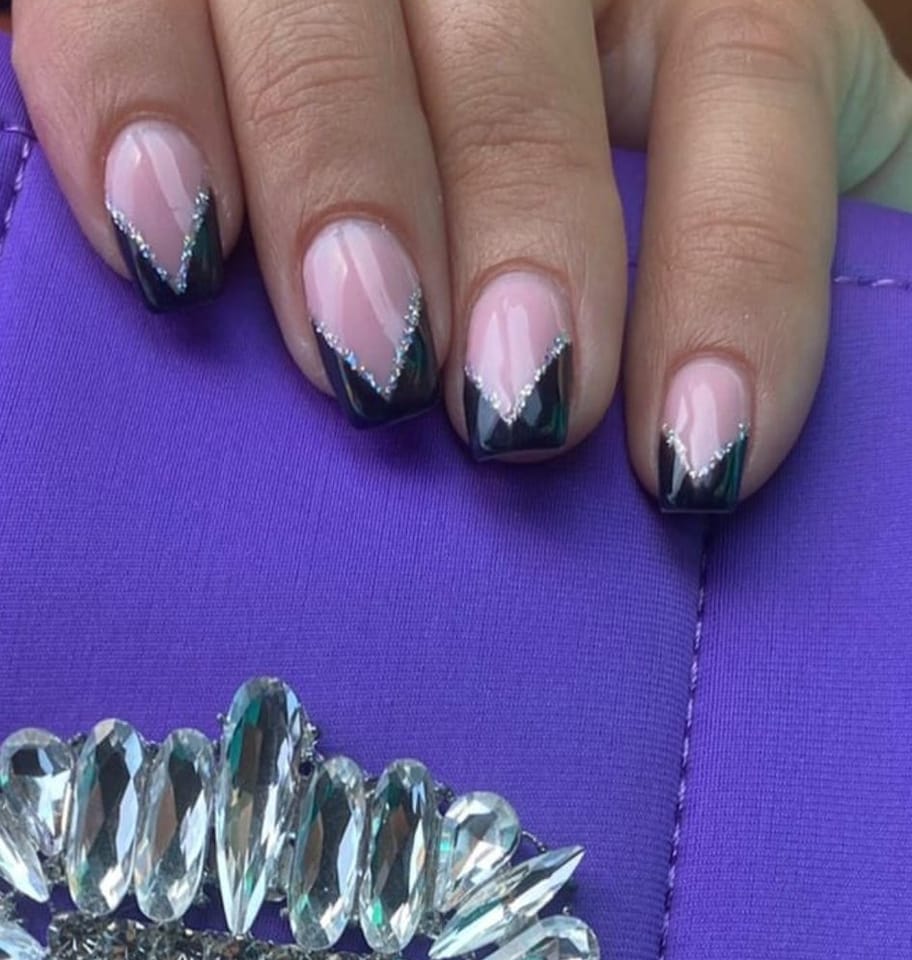 Manicura nude con puntas en V negro‑verde y borde de brillo plateado; mano sobre bolso morado con broche de cristal.